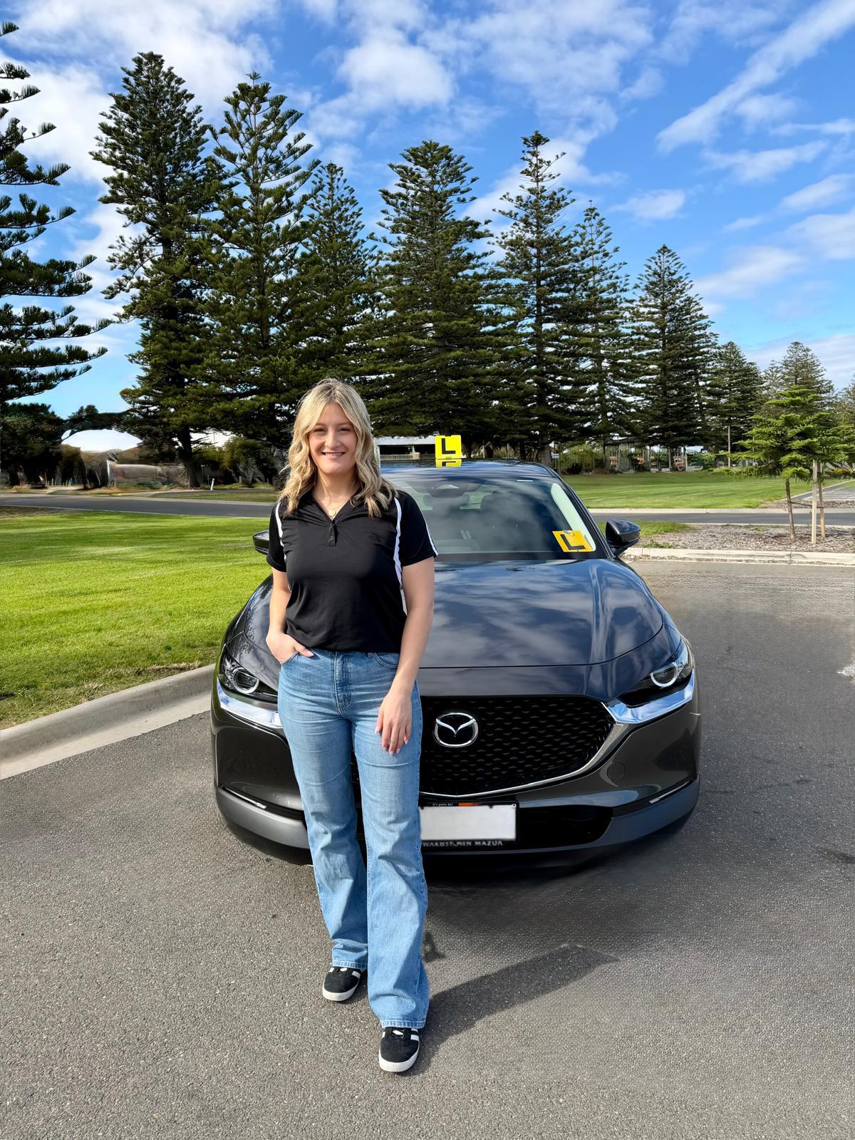 Sarah standing in front of her grey Mazda CX-30 driving lesson car fitted with L plates, beside a row of Norfolk pines